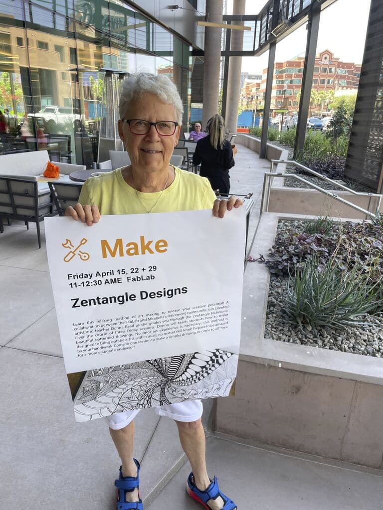A Mirabella at ASU resident standing outdoors holds up a sign advertising a class teaching how to make Zentangle Designs.