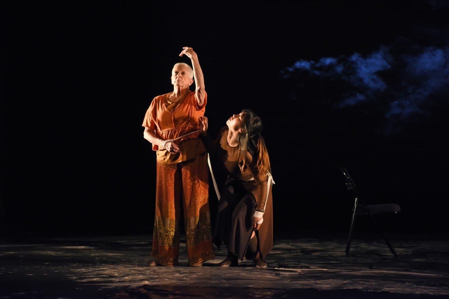 Two people perform a dance onstage under dramatic blue lighting. There is a chair to their right.